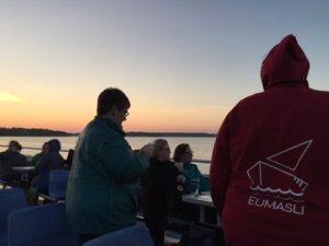 picture of the upper deck of a boat with students sitting and standing around looking away from the camera into a sunset. Student standing close to the camera is wearing a sweater with EUMASLI and a boat logo in red.